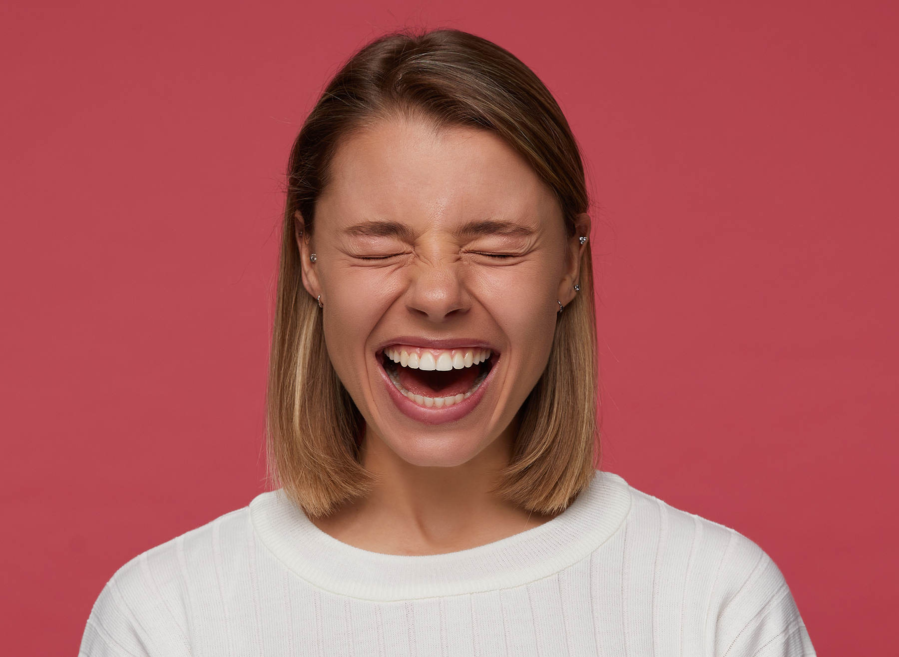 Young woman with shoulder-length blonde hair laughing with eyes closed against a red background.